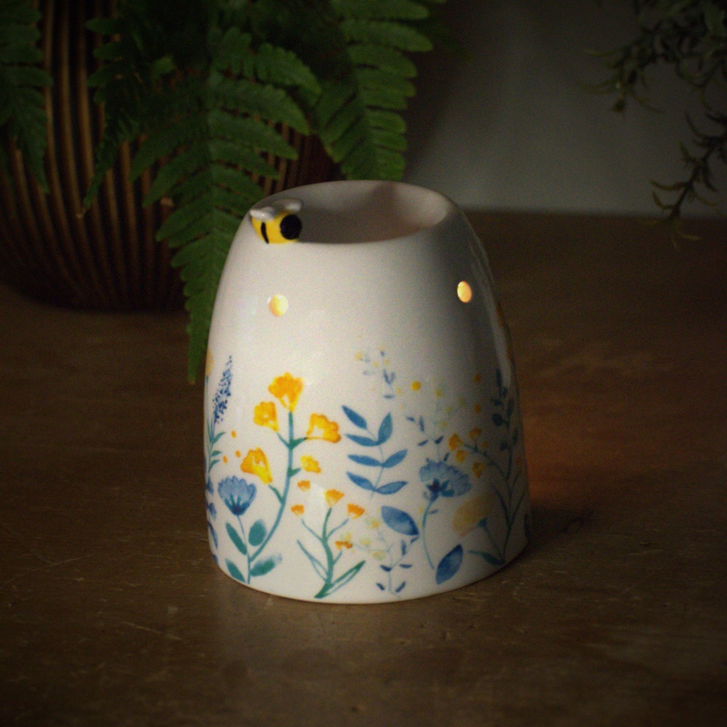 Decorative ceramic wax burner with a lit candle inside, little bumble bee on top surrounded by plants on a wooden surface.