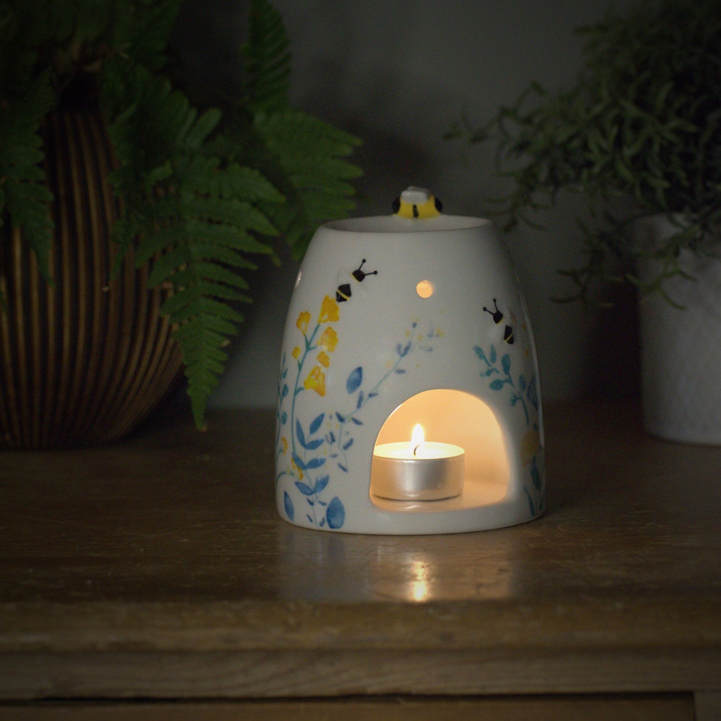 Decorative ceramic wax burner with a lit candle inside, little bumble bee on top surrounded by plants on a wooden surface.