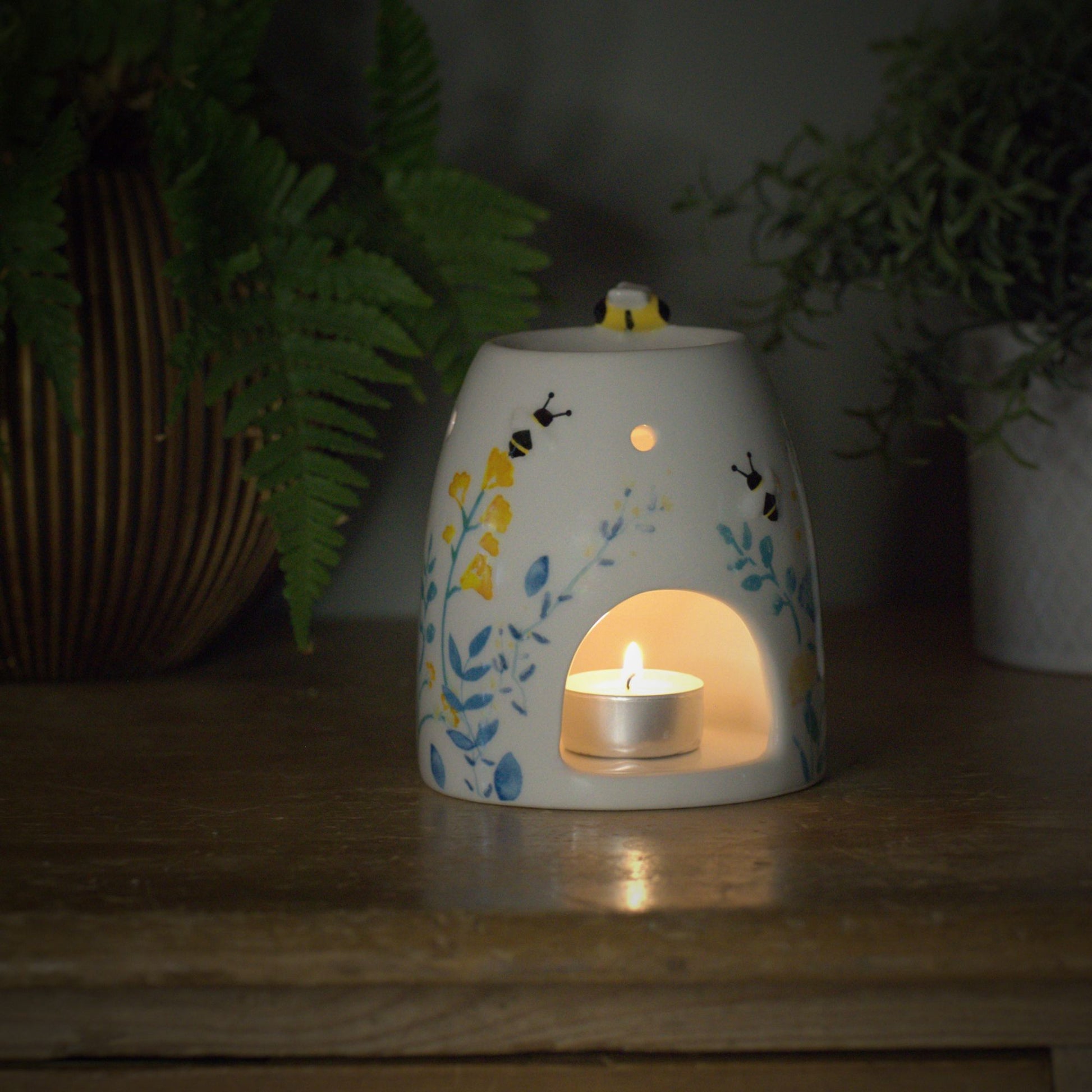Decorative ceramic wax burner with a lit candle inside, little bumble bee on top surrounded by plants on a wooden surface.