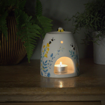 Decorative ceramic wax burner with a lit candle inside, little bumble bee on top surrounded by plants on a wooden surface.