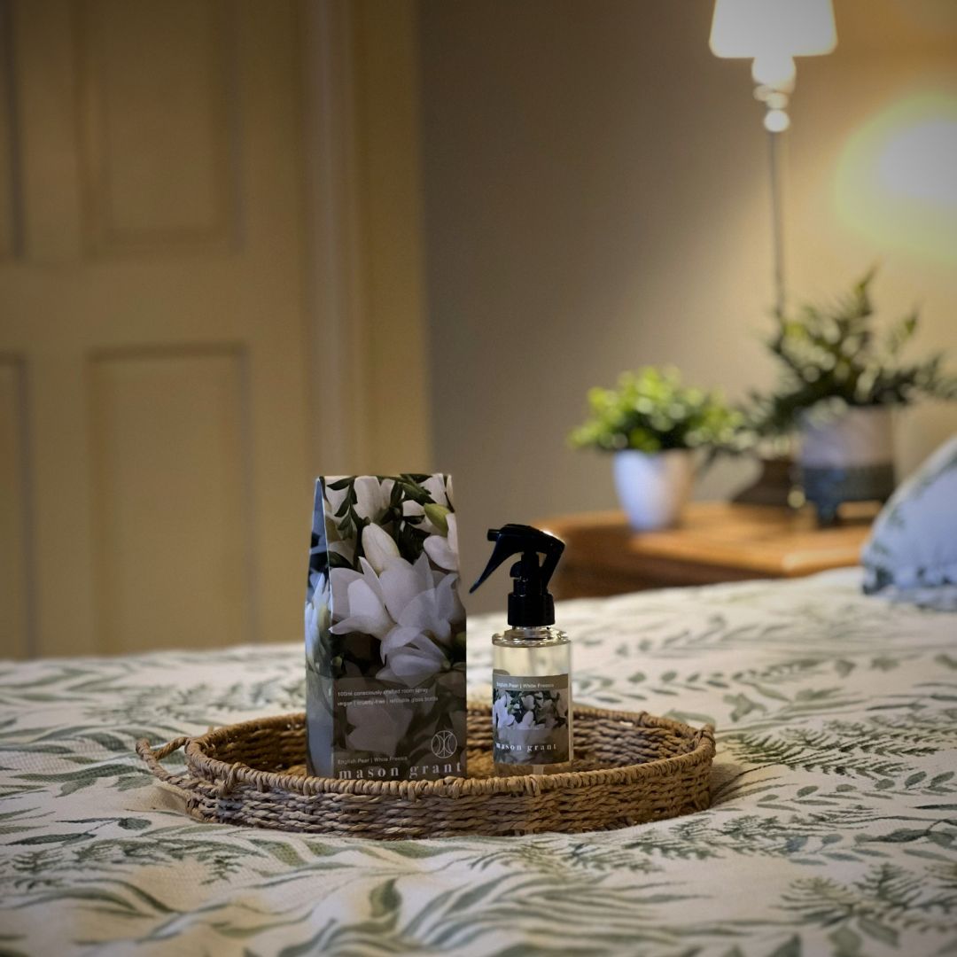 Mason Grant room spray bottle and box on wicker tray on bed with plants in the background