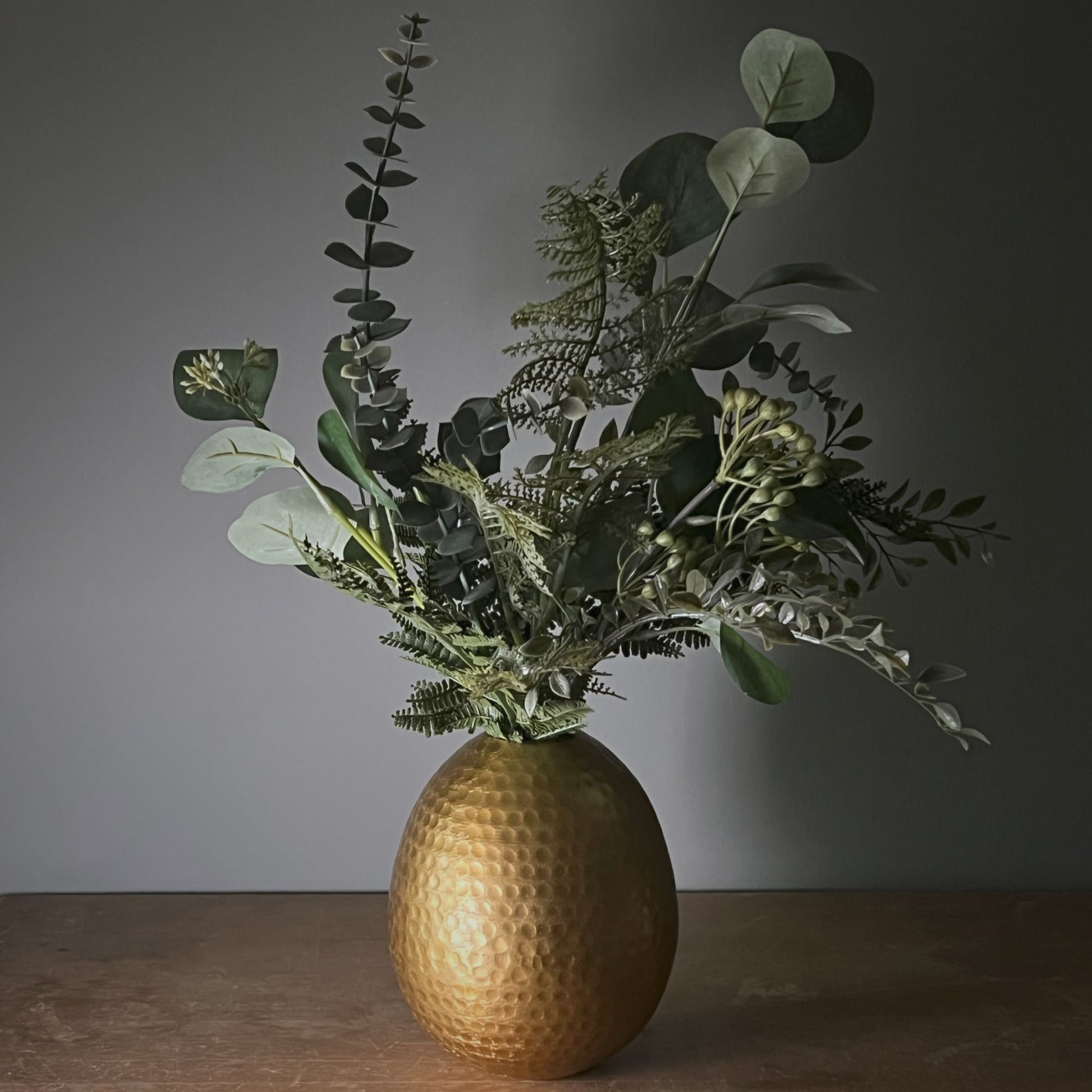 Gold textured vase with green foliage on a wooden surface against a dark background